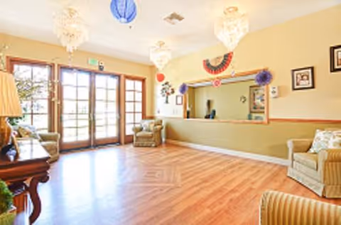 Bright reception lobby with wood floors, upholstered seating, chandeliers, and a service window near glass double doors.