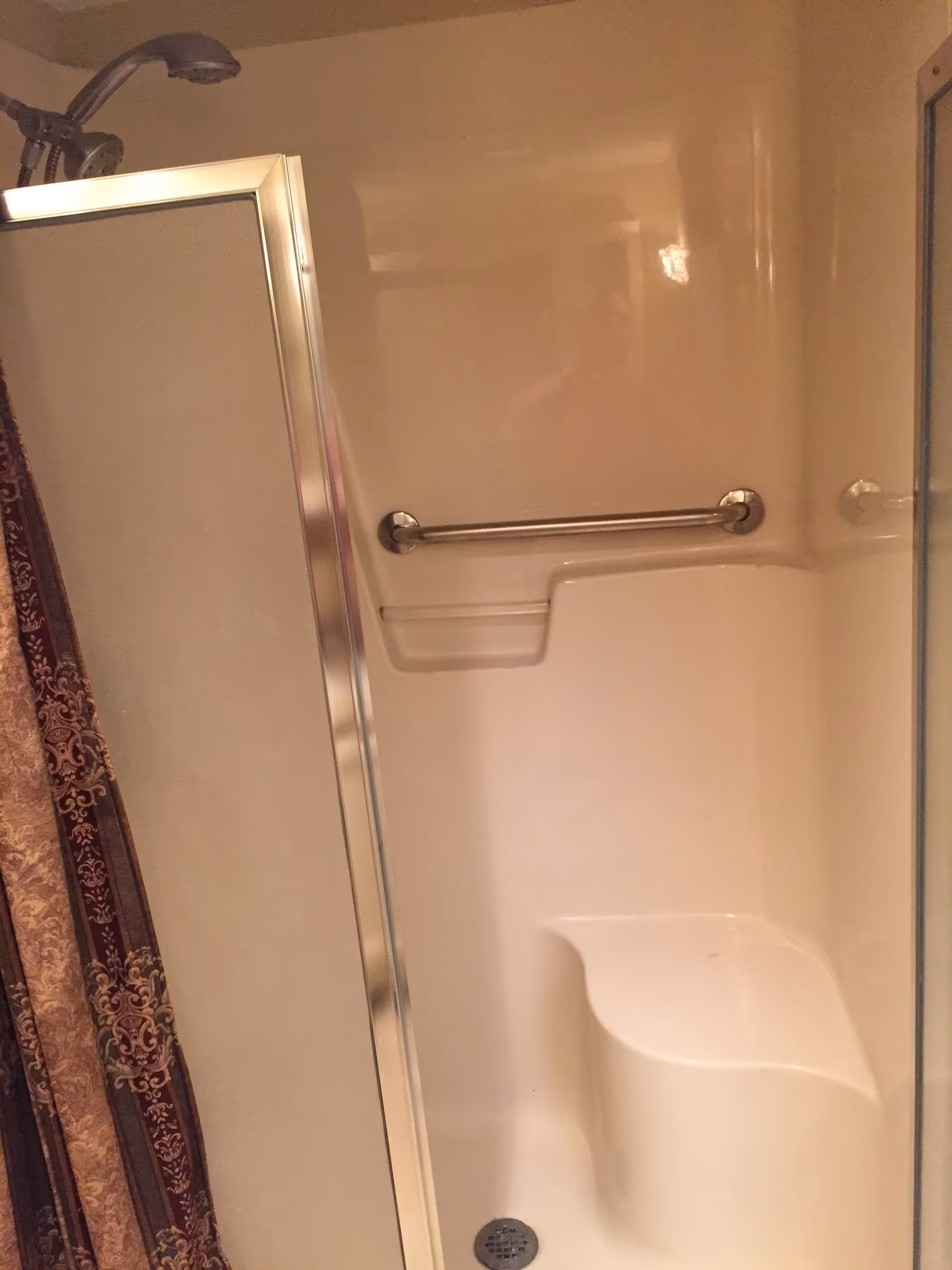 Interior view of a shower stall with a built-in seat, a grab bar on the wall, and a handheld showerhead. There is a partially visible shower door with a metal frame and a patterned shower curtain on the left side.