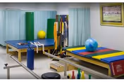A physical therapy or rehabilitation room with padded tables, exercise balls, resistance bands, and various therapy equipment. The room has light-colored walls, curtains, and a framed picture on the wall.