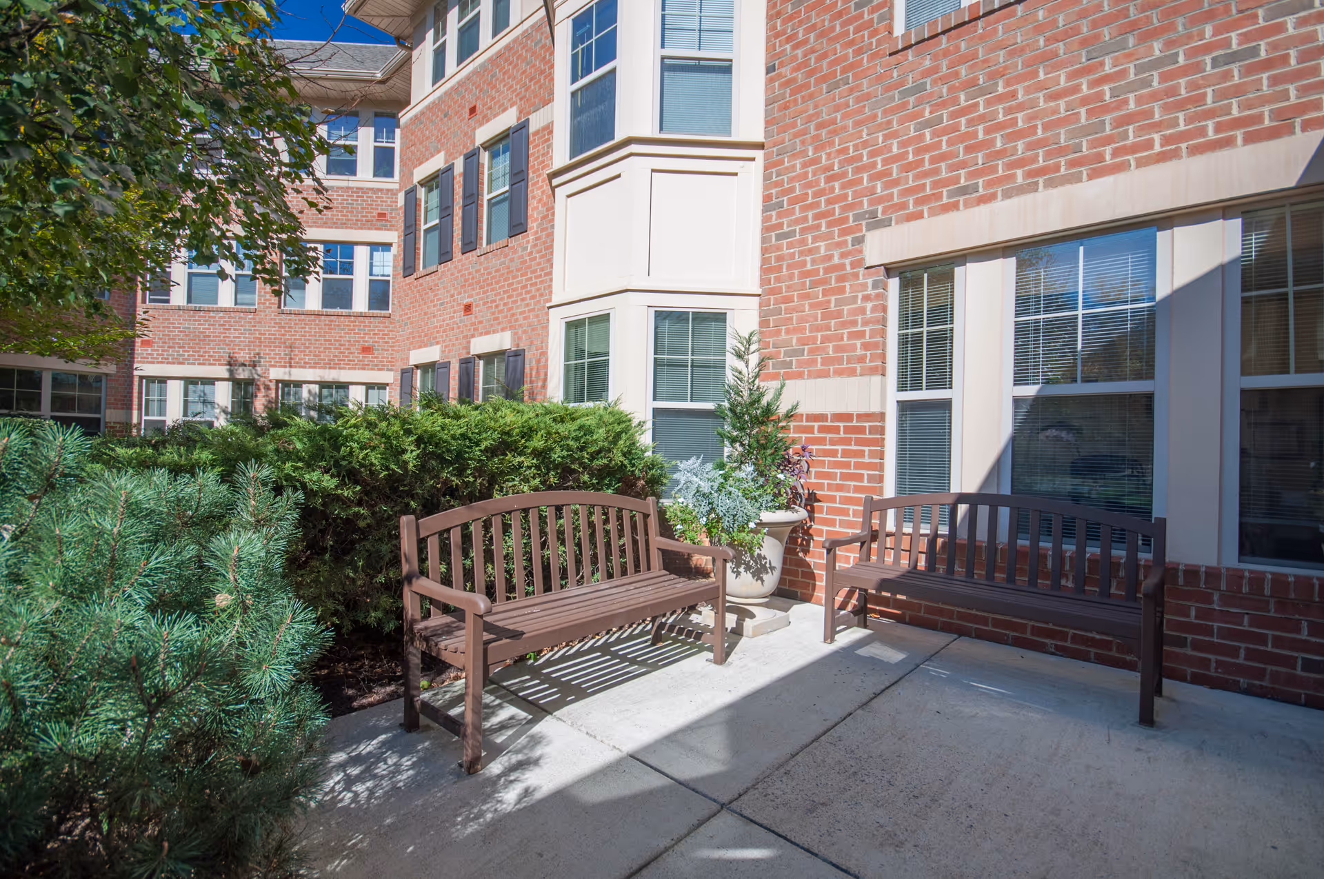 Outdoor patio area at Victory Terrace (Seniors 62+) featuring two wooden benches on a concrete surface, surrounded by green bushes and plants, with a red brick building with multiple windows in the background under a clear blue sky.
