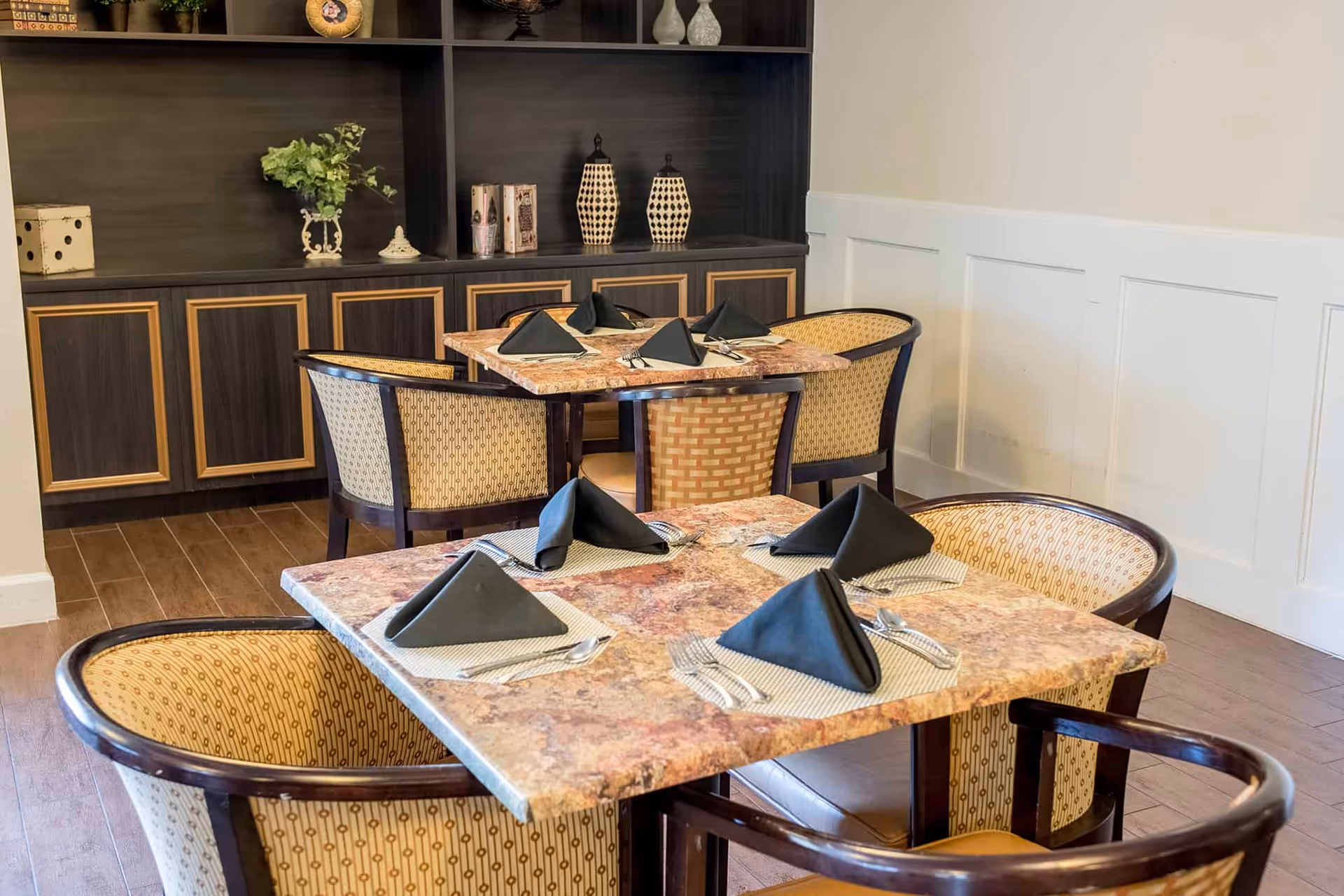 A dining area with two square tables set with black folded napkins, silverware, and placemats. The tables have a marble-like surface and are surrounded by patterned cushioned chairs. In the background, there is a dark wooden cabinet with decorative items including vases, books, and a plant.
