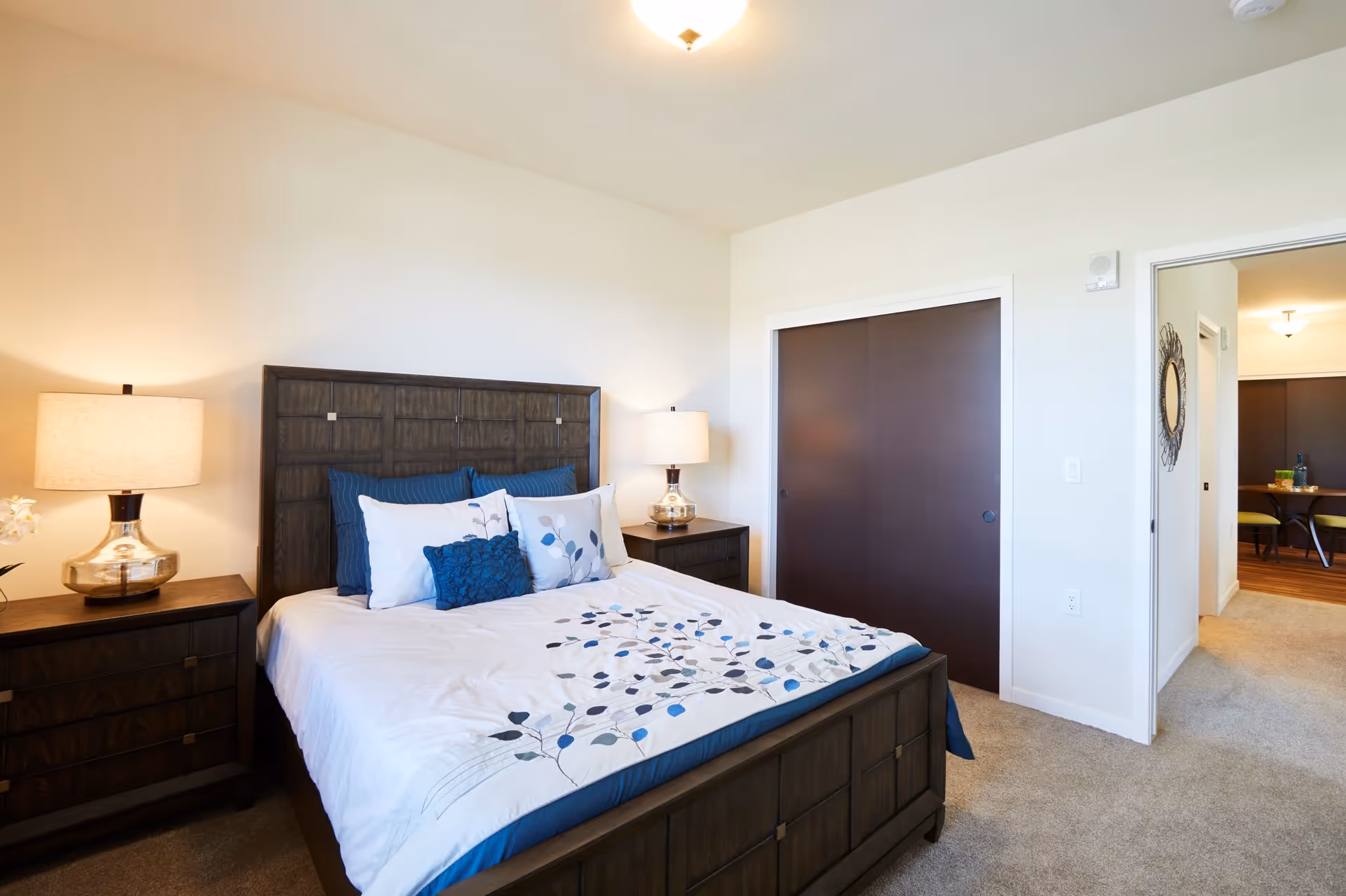 A bedroom with a dark wooden bed frame and matching nightstands on each side. The bed is made with white bedding featuring a blue and gray leaf pattern, and several pillows in white and blue. Two lamps with beige shades sit on the nightstands. There is a closet with dark sliding doors on the right wall, and an open doorway leading to another room with a dining table and chairs.