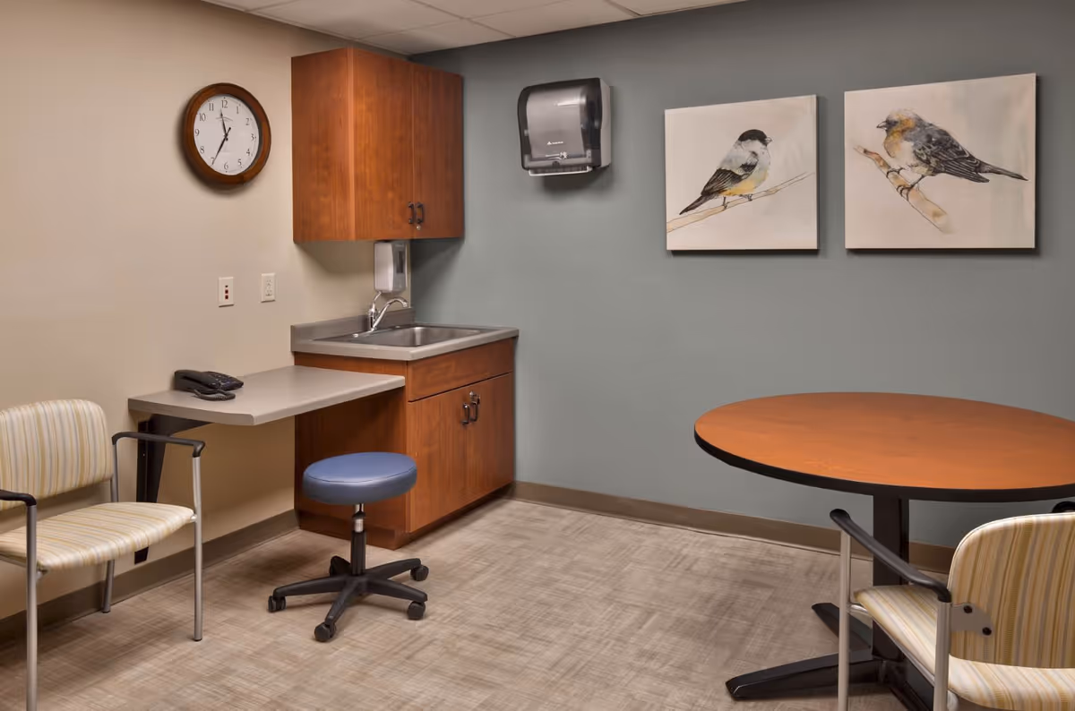 A small room with a round wooden table and two striped chairs, a wall-mounted clock, a wooden cabinet with a sink, a paper towel dispenser, and two paintings of birds on the wall.