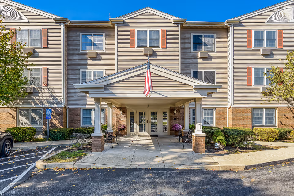Front exterior view of a three-story senior living facility with beige siding and brick accents. The entrance has a covered porch with white columns and an American flag hanging above. There are benches on either side of the entrance and some landscaping with bushes and small trees. A handicap parking sign is visible near the entrance.