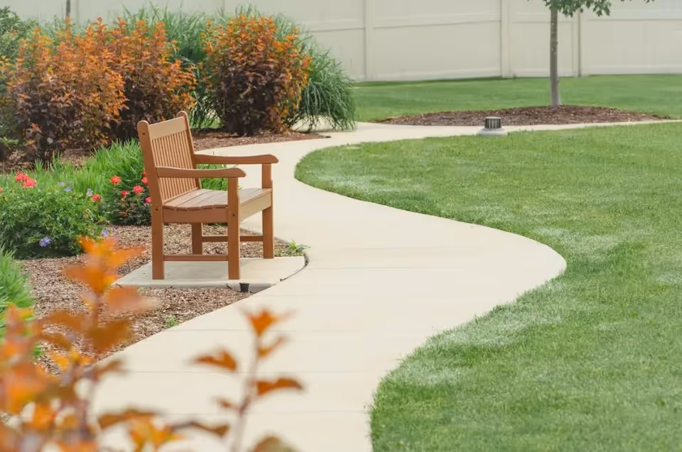 A curved concrete pathway in a garden area with a wooden bench on the left side, surrounded by green grass, bushes, and small trees, with a beige fence in the background.