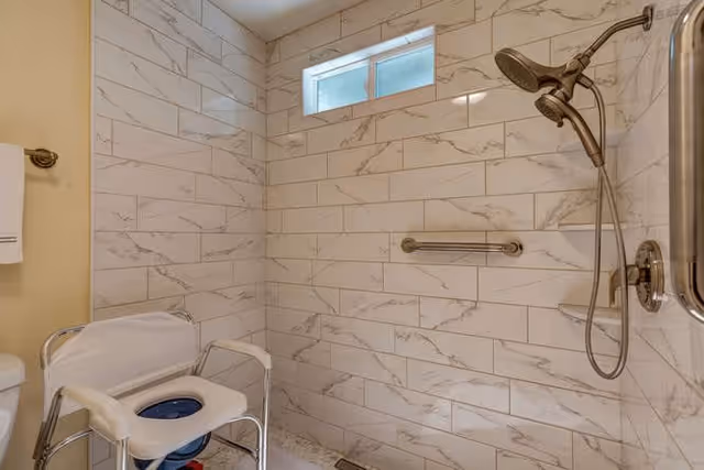 A bathroom shower area with white marble-patterned tiles on the walls, a small frosted window near the ceiling, a handheld showerhead with a hose, a grab bar, and a white shower chair with armrests positioned inside the shower space.