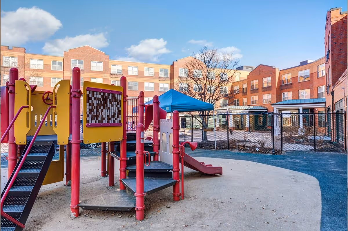 Outdoor playground area with red and yellow play structures including slides and climbing steps, set within a fenced courtyard of a multi-story brick building under a blue sky with some clouds.