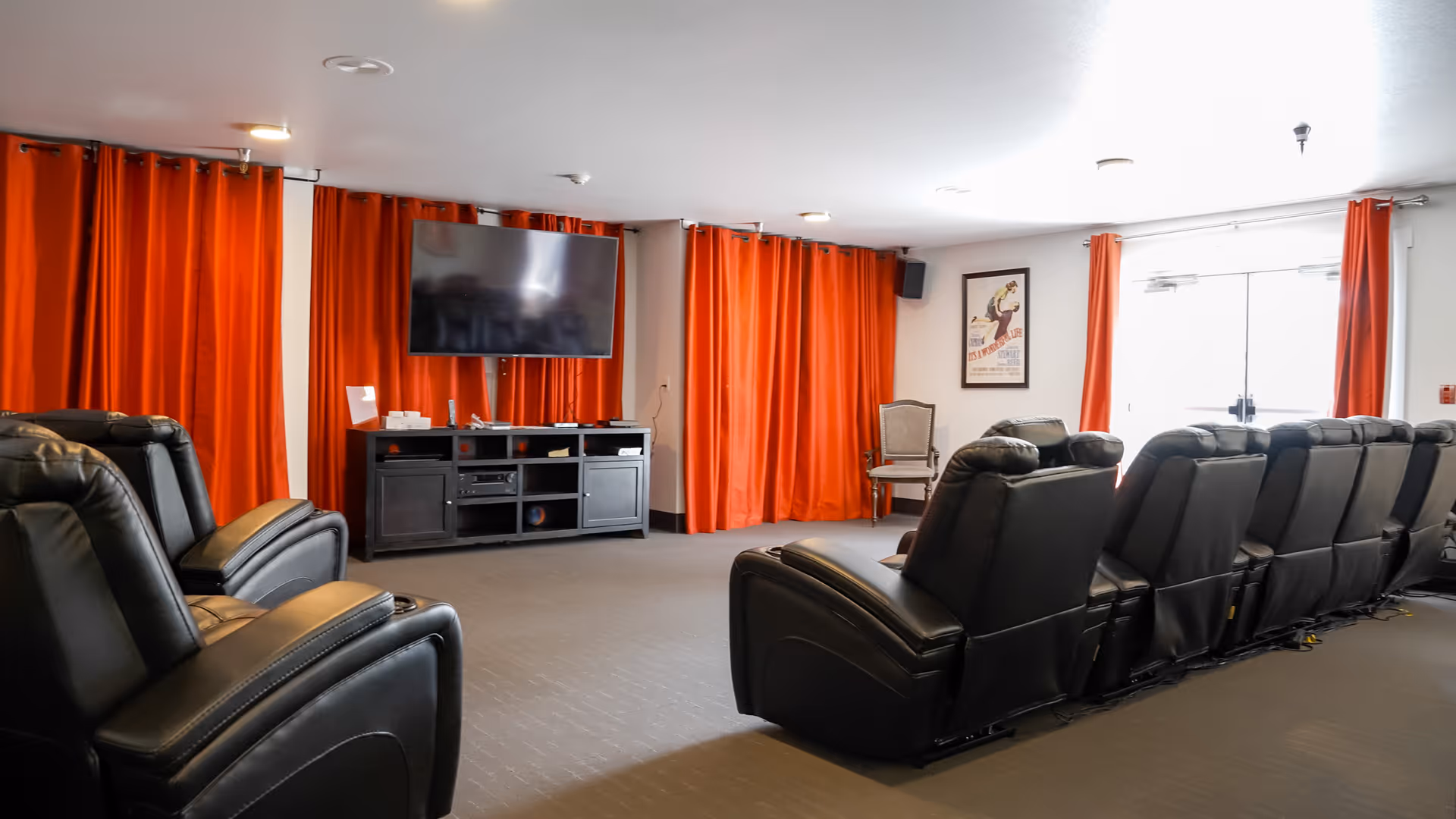 A senior living facility common room with two rows of black leather recliner chairs facing a large flat-screen TV mounted on a black entertainment unit. The room has bright orange curtains covering the windows and a framed poster on the wall. There is a single wooden chair near the wall and double glass doors letting in natural light.