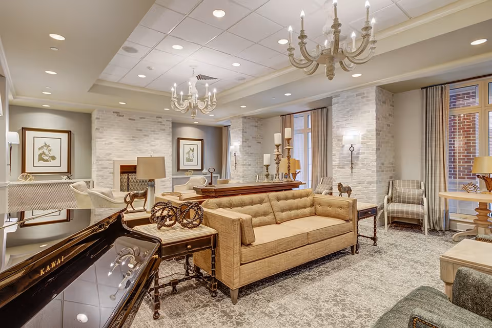 A spacious and well-lit living room in a senior living facility featuring a beige tufted sofa, a black Kawai grand piano, multiple armchairs, a wooden coffee table with decorative items, chandeliers, framed artwork on the walls, and large windows with curtains allowing natural light to enter.