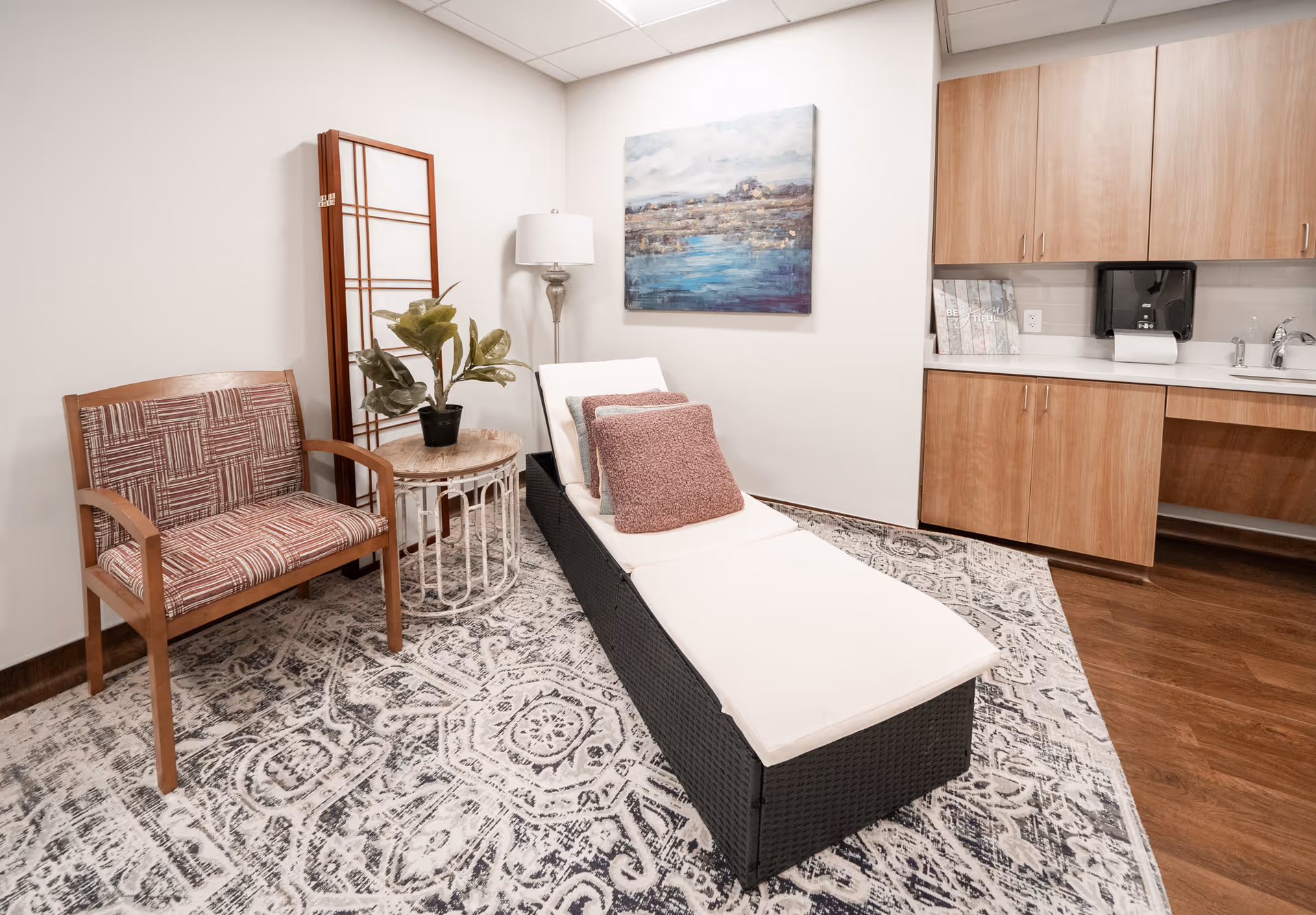 Quiet lounge-style interior with a cushioned chaise lounge, patterned armchair, side table with a plant, wall art, and wooden cabinets.