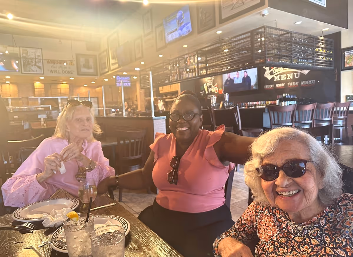 Three smiling women seated at a restaurant table with plates and drinks, with a bar and TVs visible in the background.
