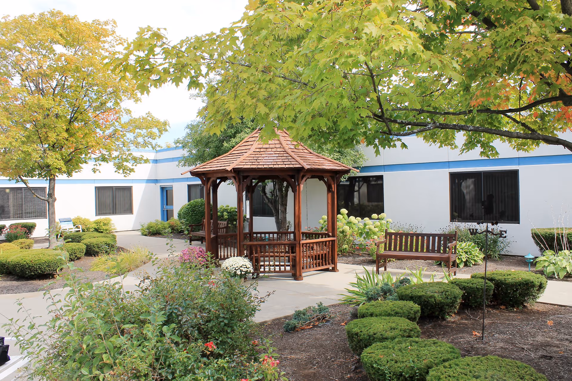 A peaceful outdoor garden area at a senior living facility featuring a wooden gazebo with benches inside, surrounded by well-maintained bushes, flowering plants, and trees with green and yellow leaves. The building with white walls and blue trim is visible in the background.