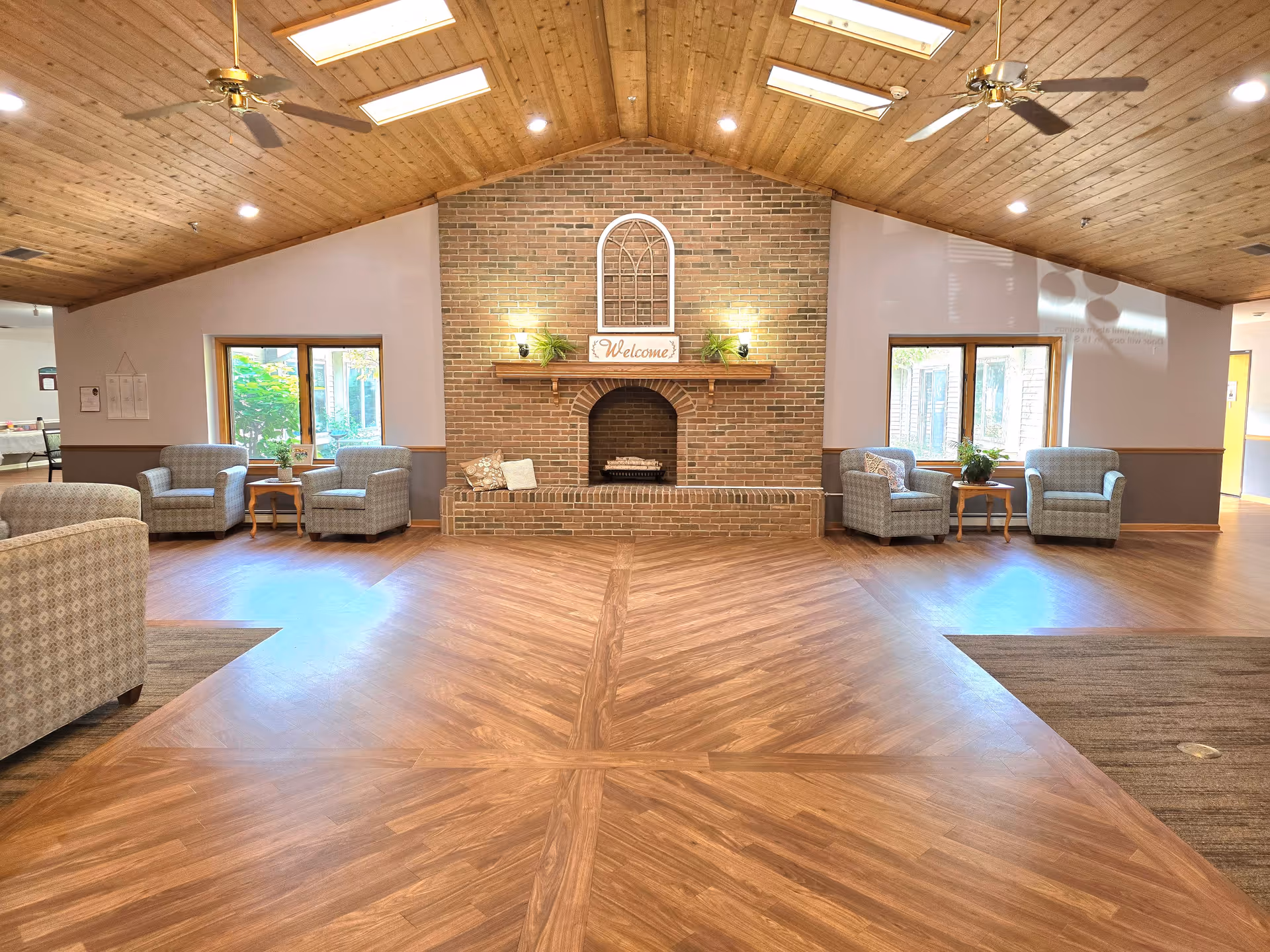 A spacious living room area with a wooden vaulted ceiling and wood-patterned flooring. The room features a central brick fireplace with a 'Welcome' sign above it, flanked by two small plants. There are two sets of armchairs and small tables positioned near windows on either side of the fireplace, allowing natural light to enter. Ceiling fans and recessed lighting are visible on the ceiling.