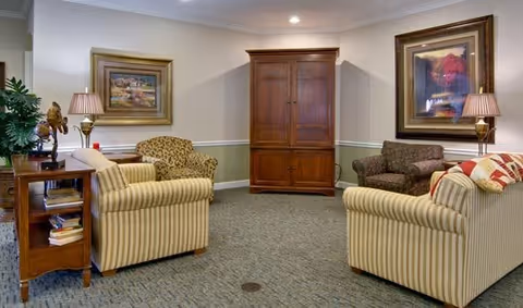 A cozy living room with two striped sofas, a patterned armchair, a wooden armoire, two table lamps with striped shades, framed artwork on the walls, and a wooden side table with books and decorative items.