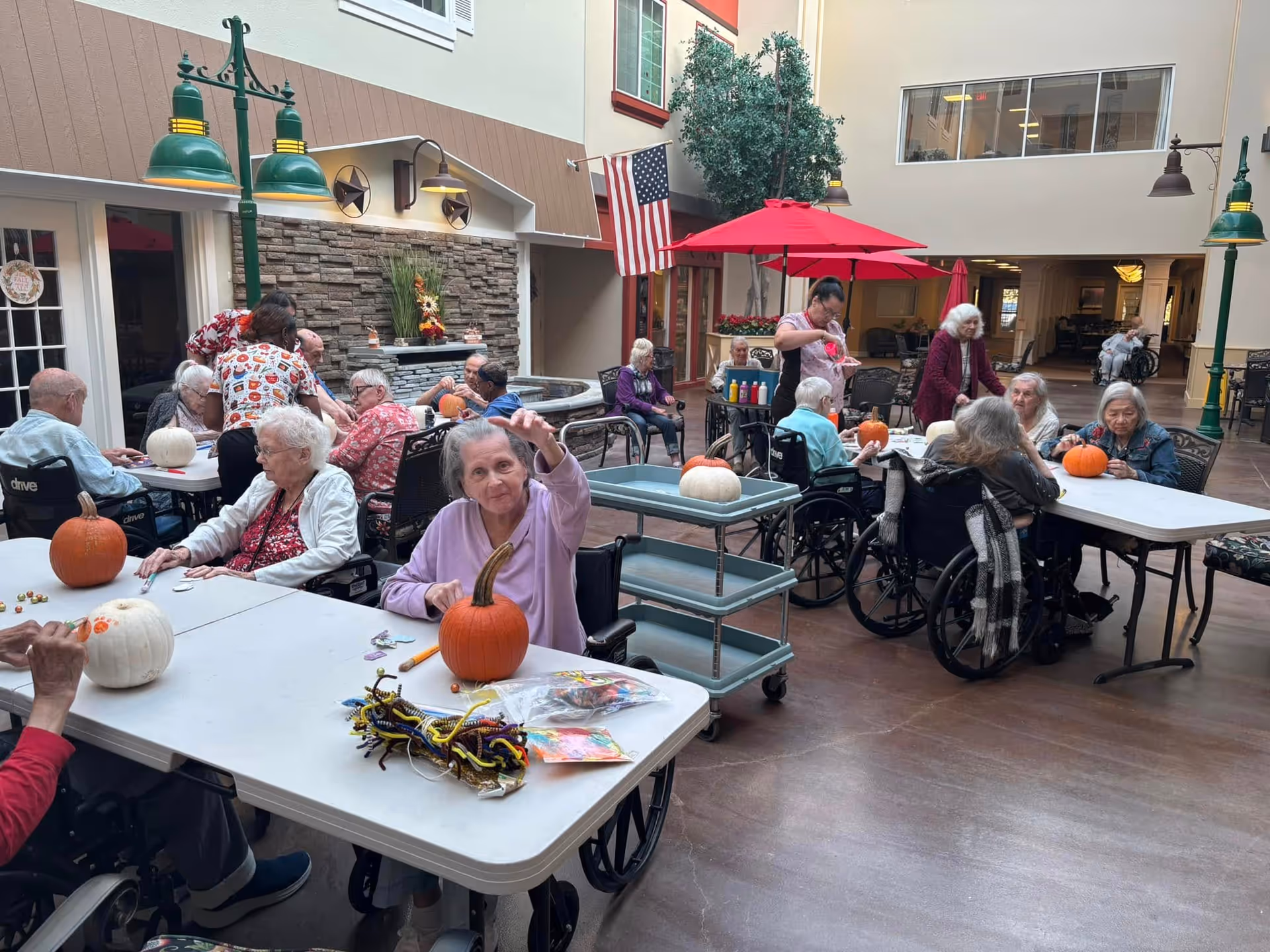 Elderly residents in wheelchairs gather at tables in an indoor common area decorating pumpkins.