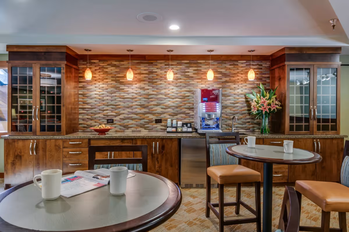 A cozy coffee and snack area with wooden cabinets, a colorful tiled backsplash, and four pendant lights hanging from the ceiling. There is a coffee machine on the counter, a bowl of snacks, and a vase with pink lilies. In the foreground, there are two round tables with chairs, each table having white mugs and one with an open magazine.