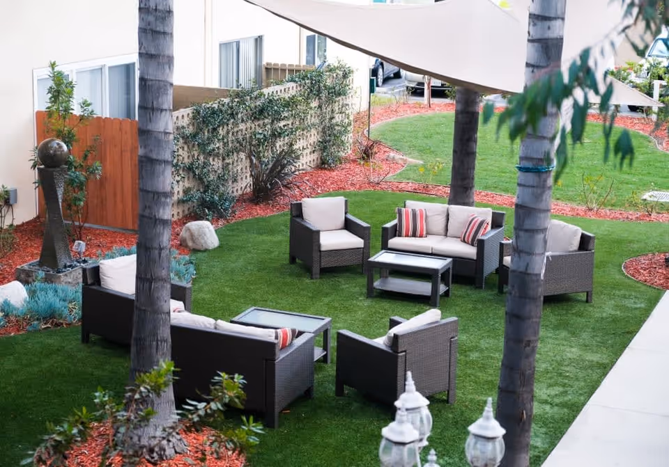 Outdoor seating area with cushioned wicker chairs and sofas arranged around glass-top coffee tables on a green lawn, surrounded by palm trees, plants, and a beige building wall with a wooden gate.