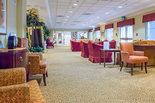 A spacious and well-lit common area with patterned carpet, multiple upholstered chairs and sofas in warm tones, side tables, potted plants, and large windows with red valances allowing natural light to fill the room.