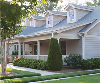 Exterior view of a single-story residential building with a covered porch, white columns, and dormer windows on the roof. The building is surrounded by green shrubs, a tree, and a well-maintained lawn with a concrete walkway.
