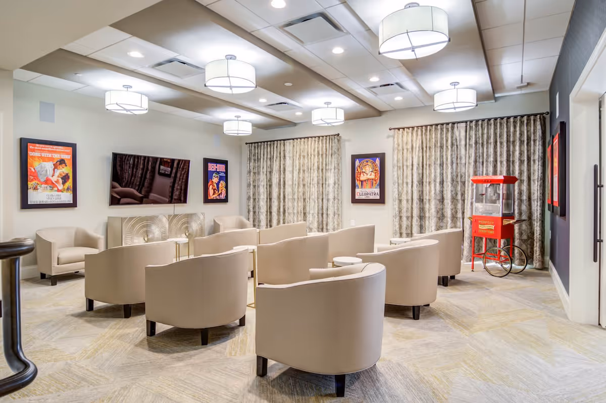 A modern common area with beige armchairs arranged in rows facing a wall-mounted flat screen TV. The room has patterned curtains, movie posters on the walls, ceiling lights, and a red vintage-style popcorn machine in the corner.