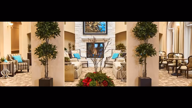 A spacious and elegant living room area with comfortable seating including sofas and armchairs adorned with blue cushions. The room features a fireplace in the center with a framed artwork above it. There are two large potted plants flanking the central seating area, and additional seating with small tables is visible on the right side near large windows with curtains. The room is warmly lit with wall sconces and has a patterned carpet.