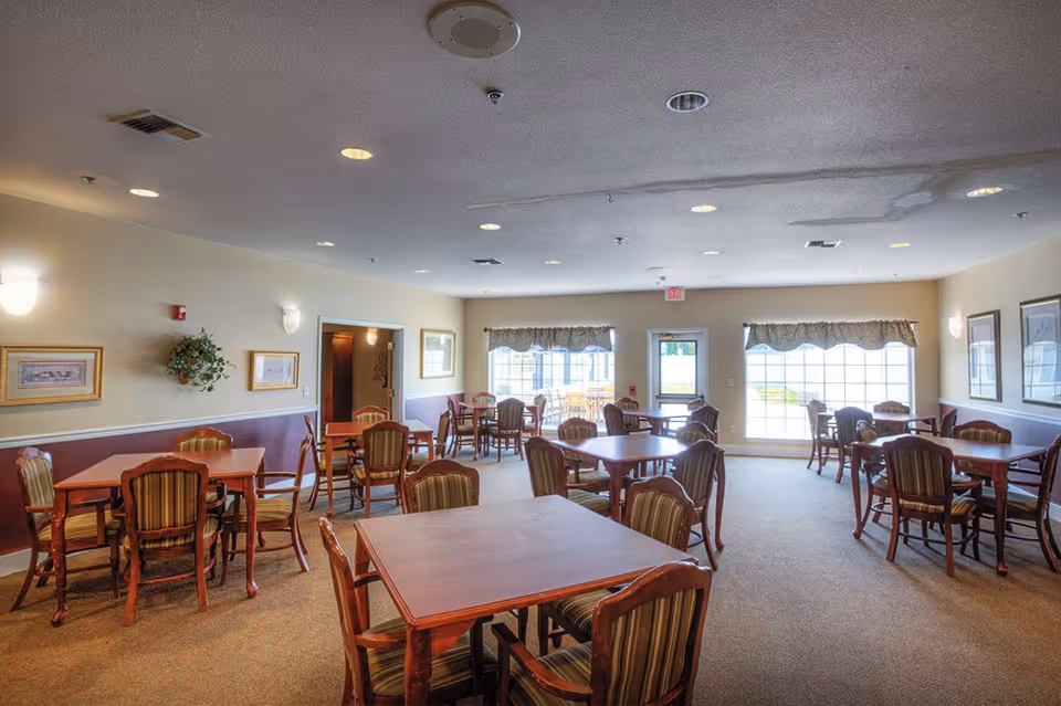 A well-lit dining room with multiple wooden tables and striped cushioned chairs arranged neatly. The room has beige walls with framed artwork and a plant on the wall. Large windows with valances allow natural light to enter, and there is a door leading outside.
