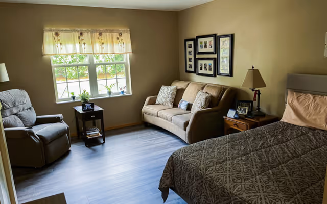 A cozy bedroom with a large bed covered in a patterned brown bedspread, a beige sofa with decorative pillows, a recliner chair, a small wooden side table with a lamp and framed photos, and a window with a floral valance letting in natural light.