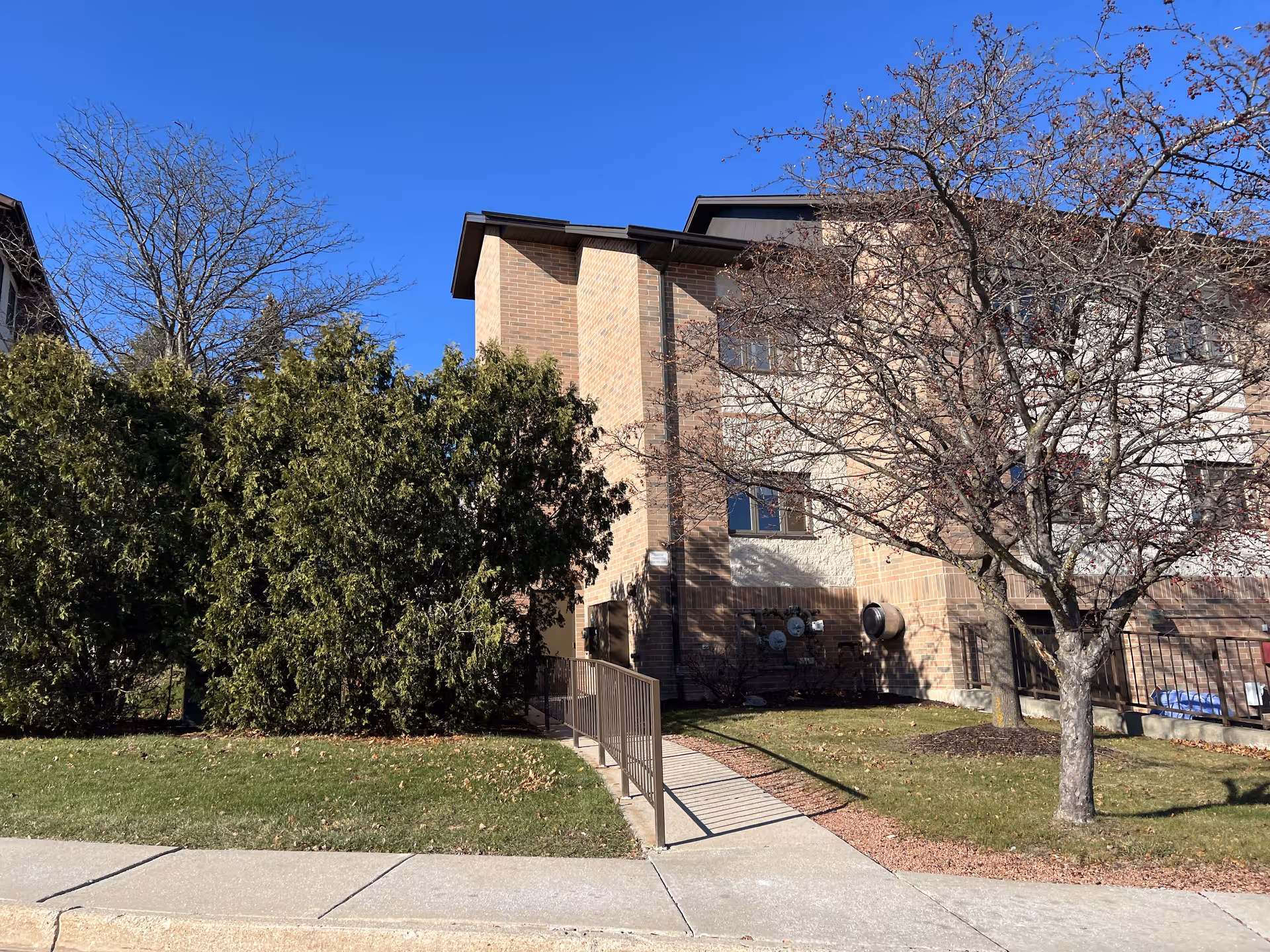 Exterior view of a multi-story brick building with a ramp leading to an entrance. There are trees and bushes around the building, and the sky is clear and blue.
