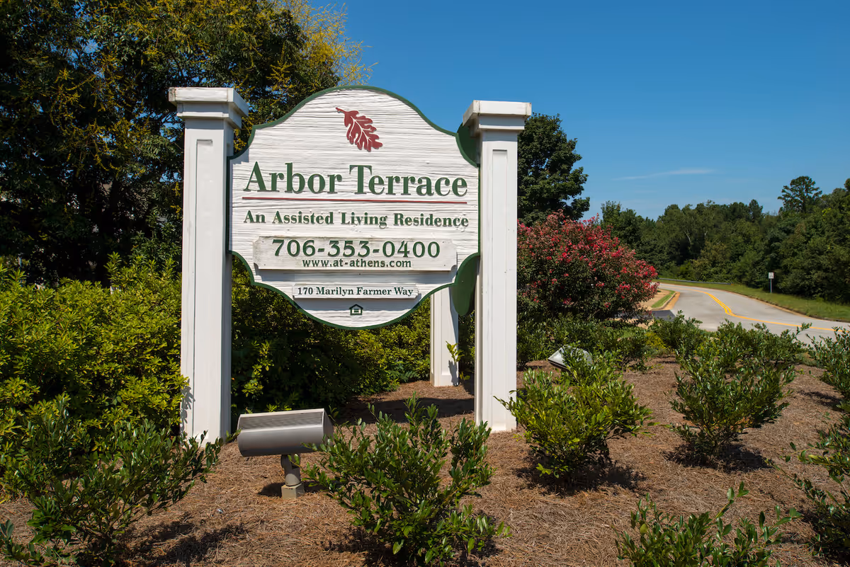 A large white wooden sign for Arbor Terrace, an assisted living residence, surrounded by green bushes and plants with a clear blue sky and a road in the background.