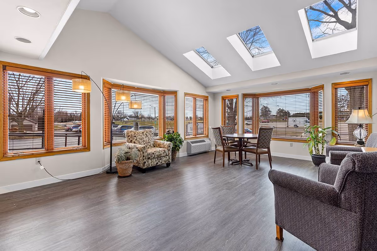 A bright and spacious senior living common area with large windows and skylights allowing natural light to fill the room. The space features a patterned armchair, a floor lamp with three lights, a round table with four chairs, and additional seating with a sofa and armchair. Several potted plants add greenery to the room, and the floor is covered with wood-like flooring.