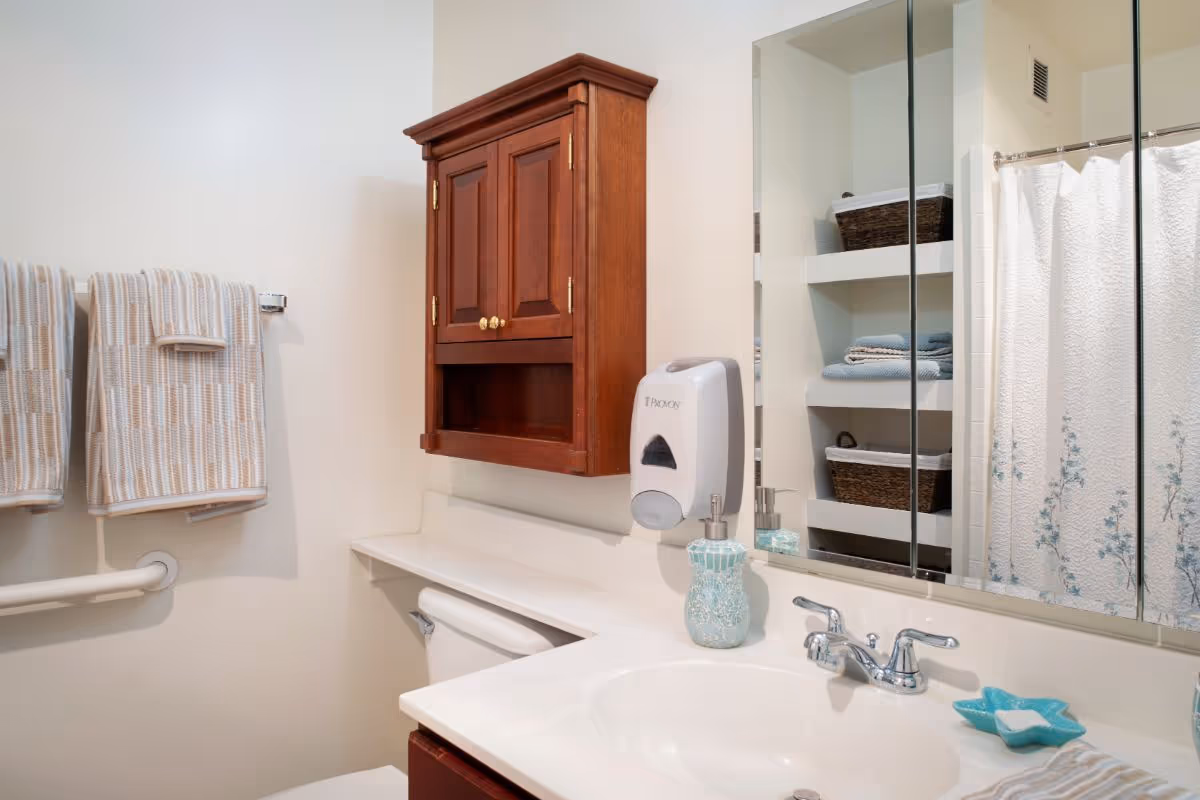 Well-lit bathroom with a sink and mirror, wooden wall cabinet, soap dispenser, folded towels, and a shower with a patterned curtain.