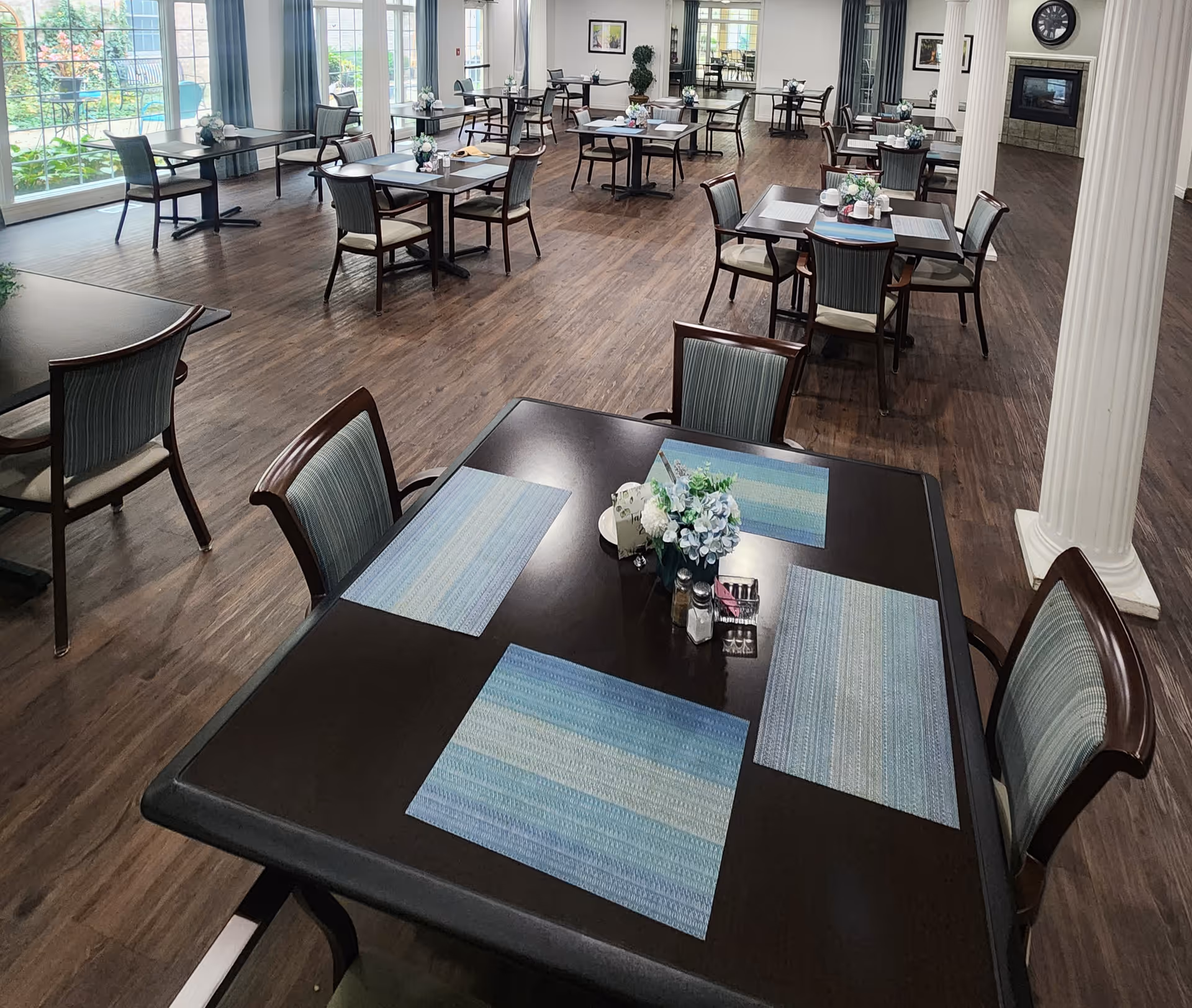 A spacious dining room with multiple dark wood tables and chairs arranged neatly. Each table has blue placemats and a small floral centerpiece. Large windows on one side let in natural light, and there is a fireplace with a clock above it on the far wall. The floor is wooden, and the room has white columns and light-colored walls.