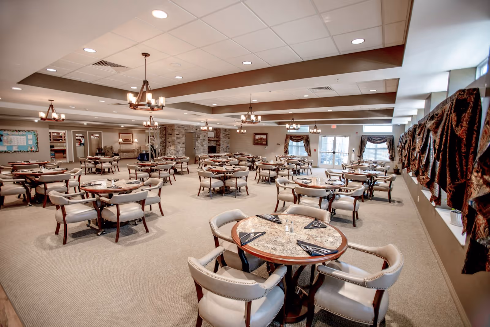 A spacious dining room with multiple round tables, each set with black napkins and silverware. The room features beige carpet, cushioned chairs with wooden legs, chandeliers hanging from the ceiling, and windows with patterned curtains on the right side. The far end of the room has a stone accent wall and a door leading outside.