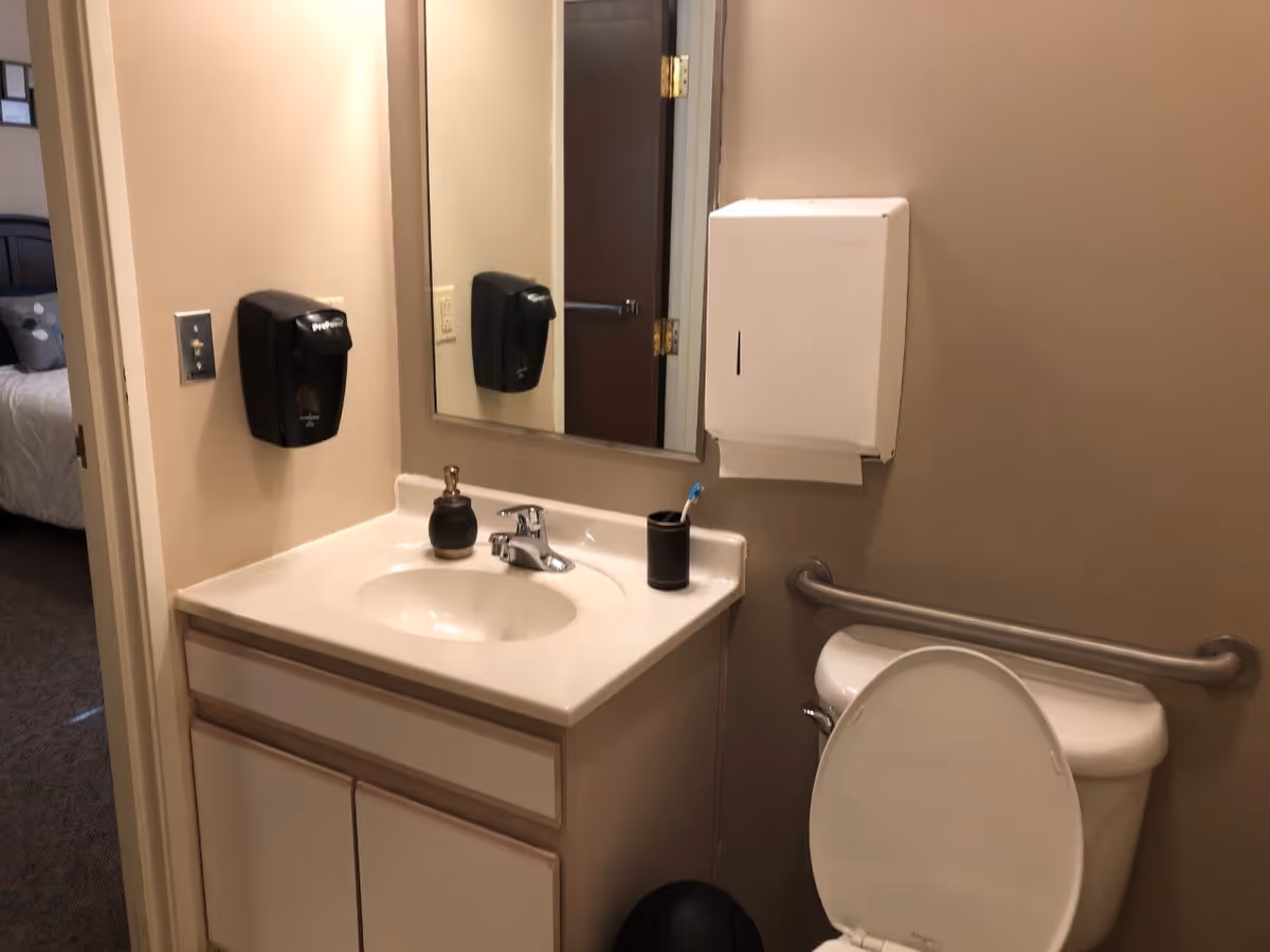 A small bathroom with a white sink and countertop, a mirror above the sink, a black soap dispenser mounted on the wall, a toothbrush holder with a toothbrush, a white paper towel dispenser, and a toilet with the lid open. A grab bar is installed on the wall next to the toilet. The bathroom door is open, showing a glimpse of a bedroom with a bed and pillows.