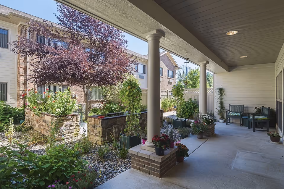 Covered outdoor patio area with white columns and brick bases, surrounded by various potted plants and flowers. Beyond the patio, there is a garden with raised stone flower beds, a tree with purple leaves, and a building with windows in the background under a clear blue sky.