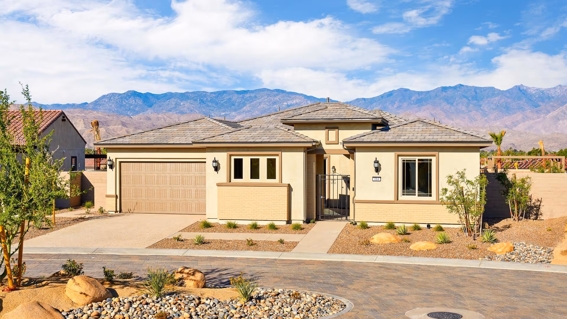 Single-story beige house with a tiled roof, a two-car garage, and a small gated front entrance. The house is surrounded by desert landscaping with rocks, small plants, and a paved driveway. Mountains are visible in the background under a partly cloudy sky.