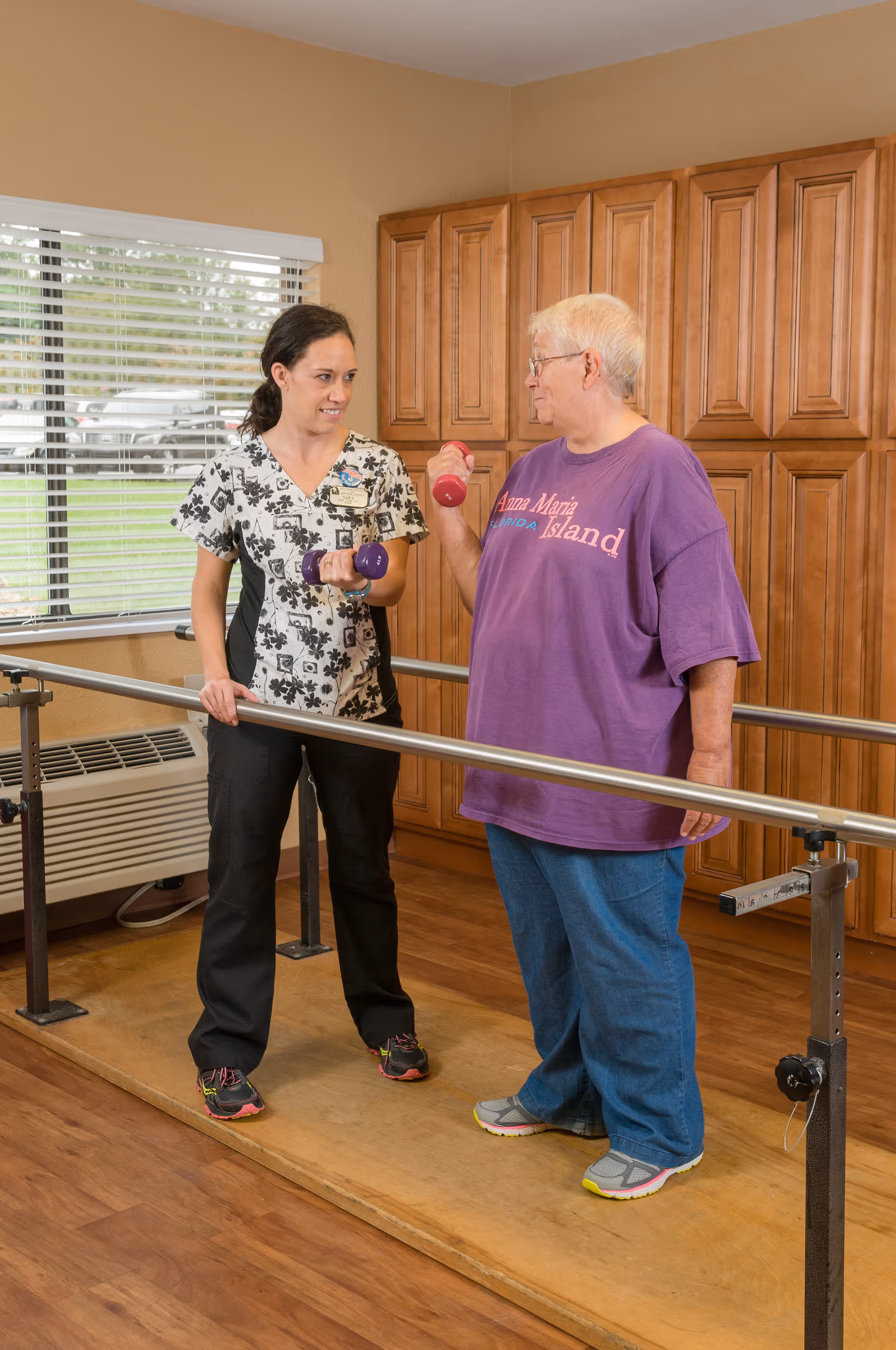 A healthcare worker assists an elderly woman with light exercise using small dumbbells in a room with wooden cabinets and a window with blinds. They are standing on a wooden platform with parallel bars for support.