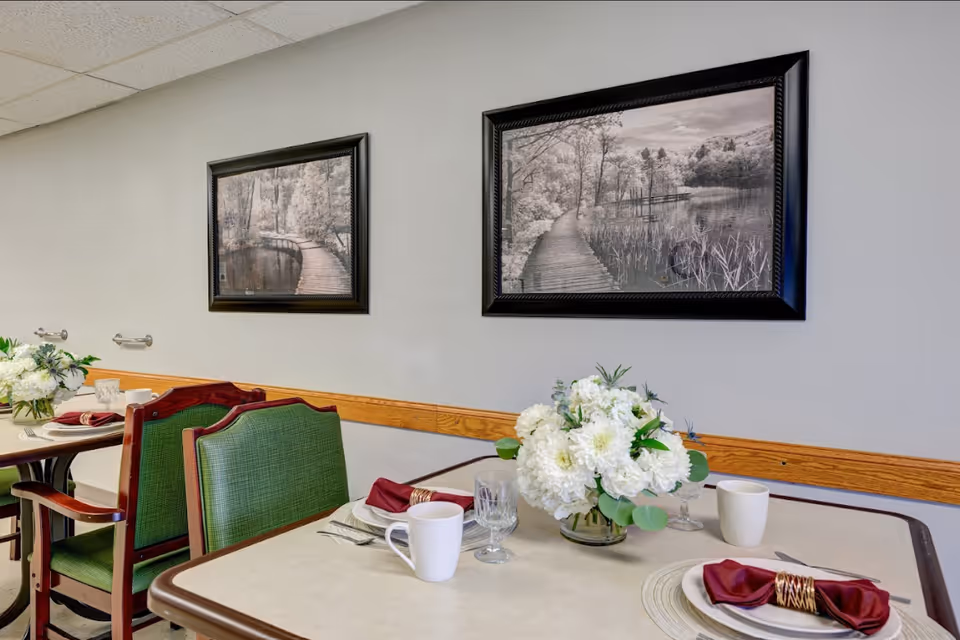 A dining area with tables set for a meal, featuring white plates, maroon napkins with gold napkin rings, white mugs, and clear glasses. Each table has a centerpiece of white flowers in a glass vase. The chairs have green upholstery with wooden frames. Two framed black and white landscape photographs hang on the light gray wall above the tables.