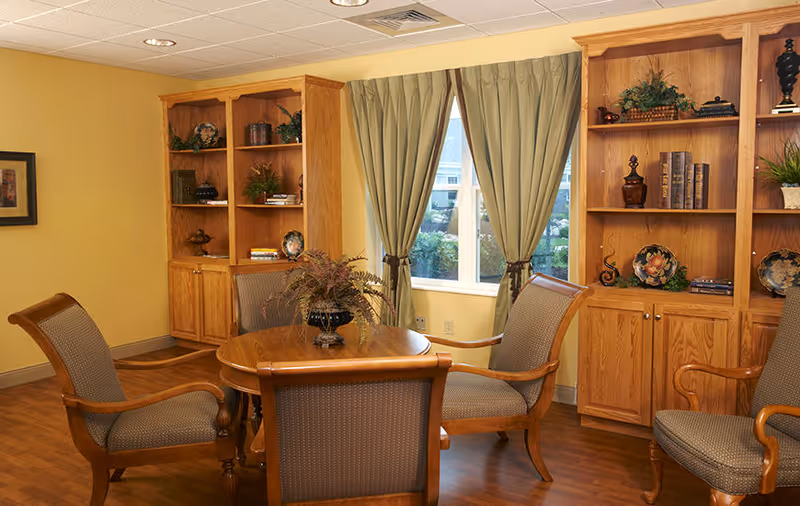 A cozy interior room with a round wooden table surrounded by four cushioned wooden chairs. Behind the table are two wooden bookshelves filled with decorative items, books, and plants. A window with green curtains lets in natural light, and the walls are painted a warm yellow color.