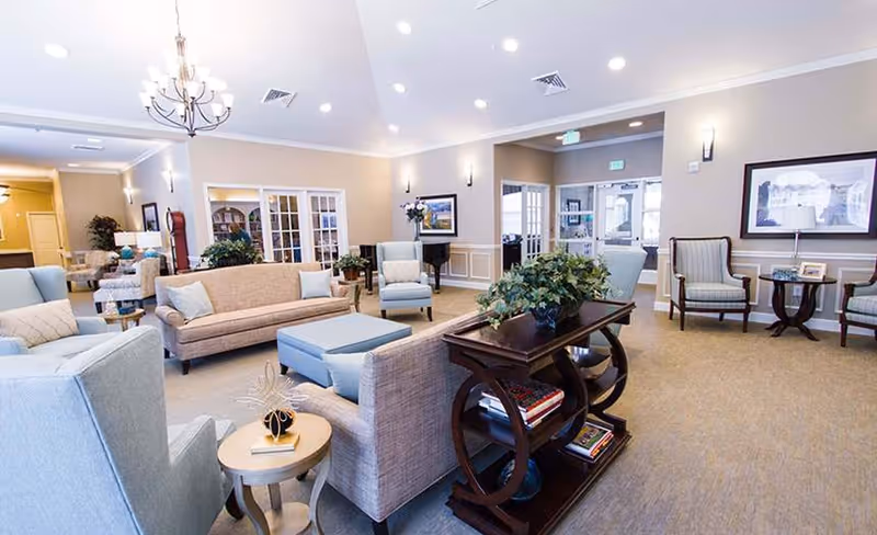 Bright, spacious common living room with sofas, armchairs, side tables, a chandelier, and decorative plants.