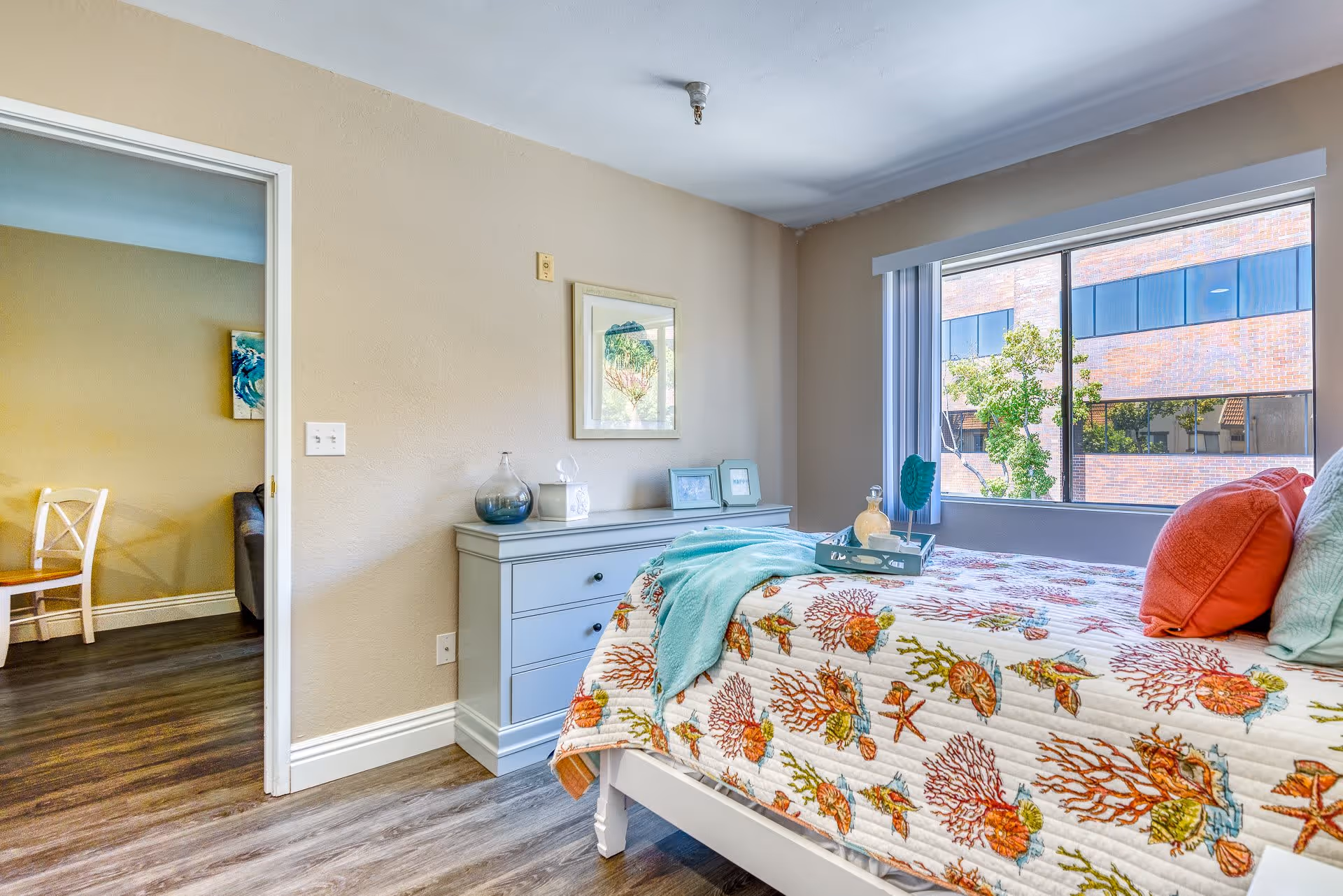 A bright bedroom in Santa Rosa Hills Senior Living featuring a bed with a colorful sea-themed quilt and coral and aqua pillows. A light blue dresser with decorative items and framed pictures sits against the beige wall. A large window with vertical blinds lets in natural light and shows a view of a brick building and trees outside. The doorway opens to another room with a wooden chair and part of a sofa visible.