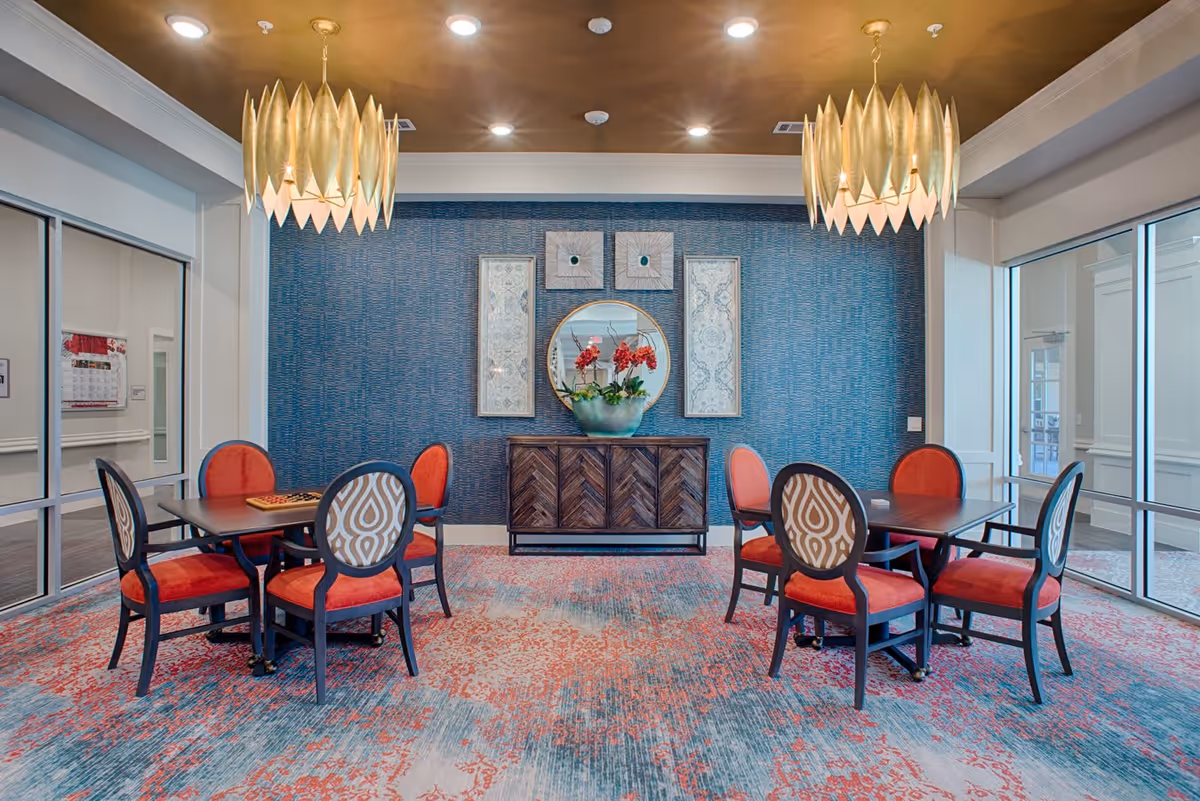 A stylish interior room with two square tables, each surrounded by four chairs with red cushions and patterned backs. The room features a blue textured accent wall with a round mirror and three decorative wall hangings above a wooden sideboard. Two large, gold pendant lights hang from the ceiling, and the floor is covered with a multicolored carpet in shades of red, blue, and beige. Glass walls and doors are visible on either side of the room.