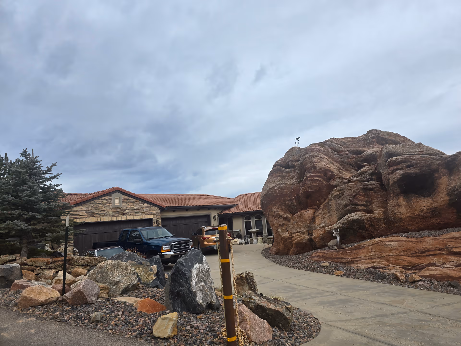 Exterior view of a single-story building with a red tile roof and three garage doors. There are two vehicles parked in front of the garages. A large rock formation is on the right side of the driveway, with a small deer statue placed near its base. The sky is cloudy and overcast.