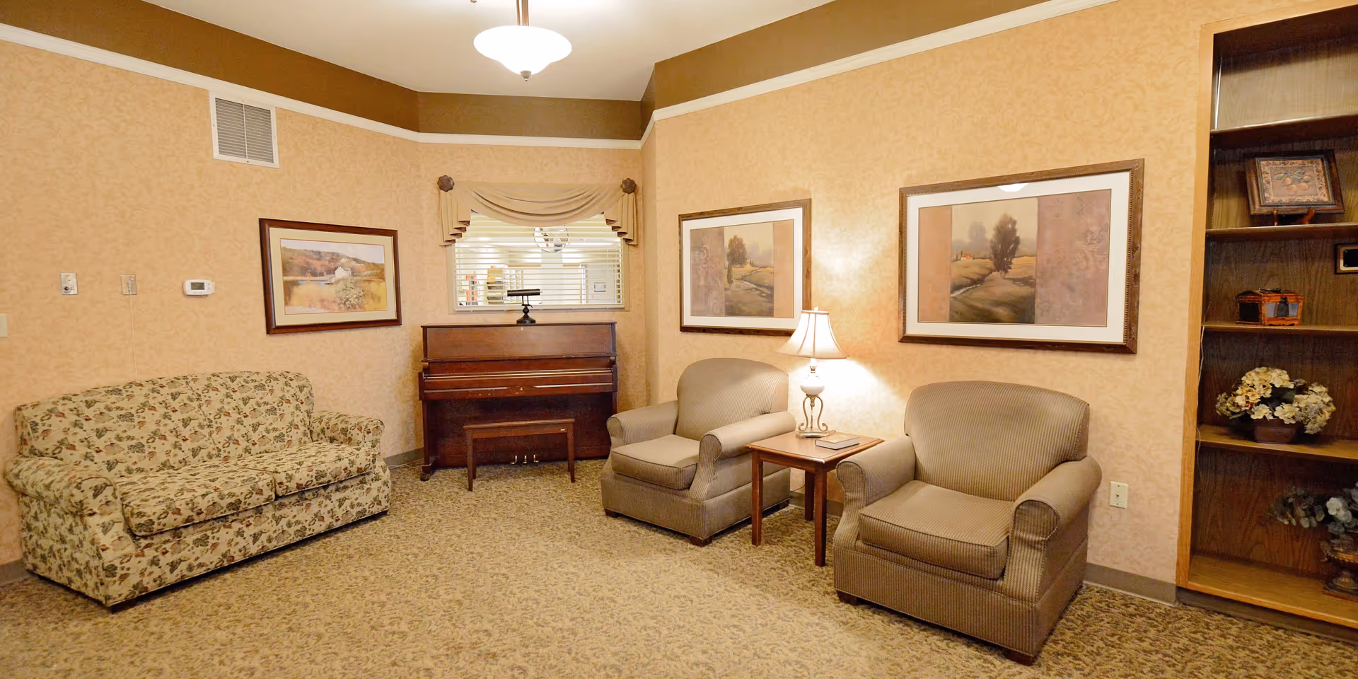 Cozy communal sitting room with a floral sofa, two armchairs, a piano, framed landscape artwork and a table lamp.