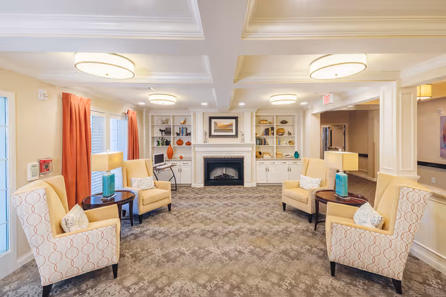 A cozy living room area in a senior living facility with four beige armchairs arranged around two small round tables with lamps. The room features a fireplace with built-in shelves on either side, decorated with books and ornaments. The walls are light-colored, and there are large windows with red curtains allowing natural light to enter.