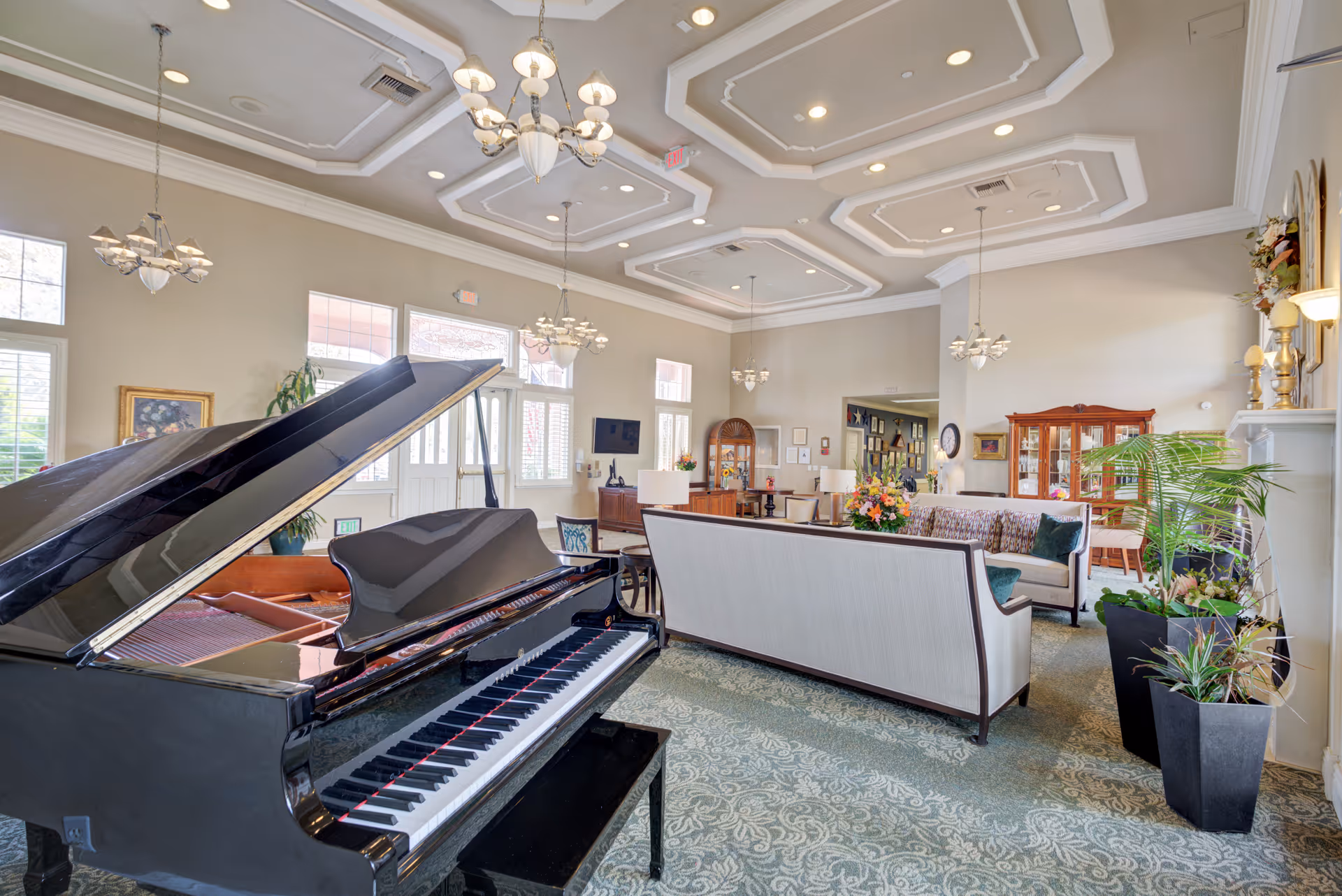 Spacious and elegant living room area with a grand piano in the foreground, multiple chandeliers hanging from a decorative ceiling, comfortable sofas arranged around a coffee table with floral arrangements, large windows allowing natural light, and various plants and decorative furniture pieces.