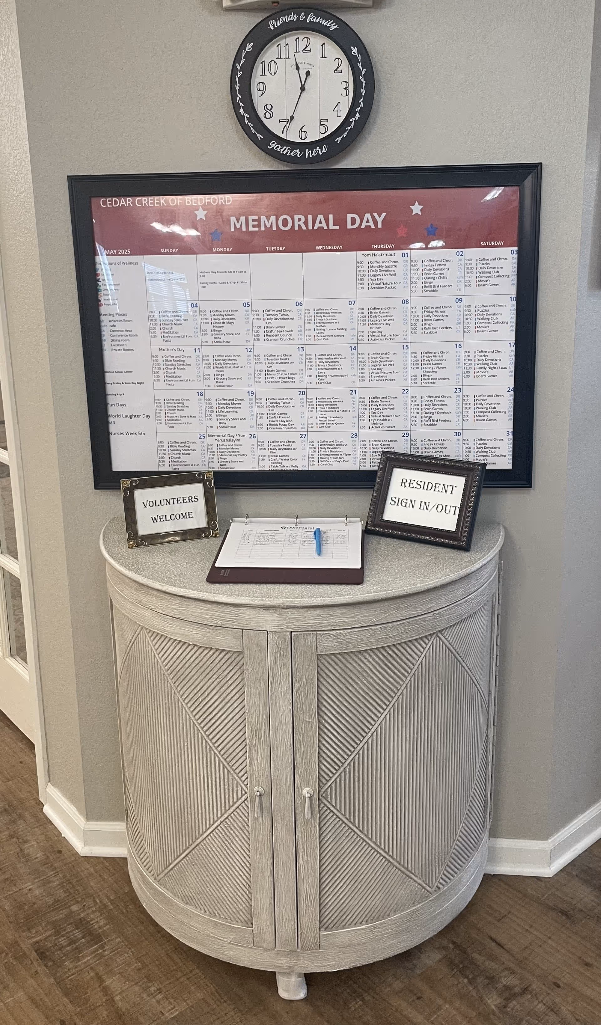 A corner area inside Cedar Creek of Bedford featuring a round wooden cabinet with a textured front. On top of the cabinet are two small framed signs reading 'VOLUNTEERS WELCOME' and 'RESIDENT SIGN IN/OUT' along with a sign-in sheet and a blue pen. Above the cabinet is a large framed calendar for May 2025 with a Memorial Day header. A round wall clock with the words 'friends & family gather here' is mounted above the calendar. The floor is wooden, and the walls are painted light gray.