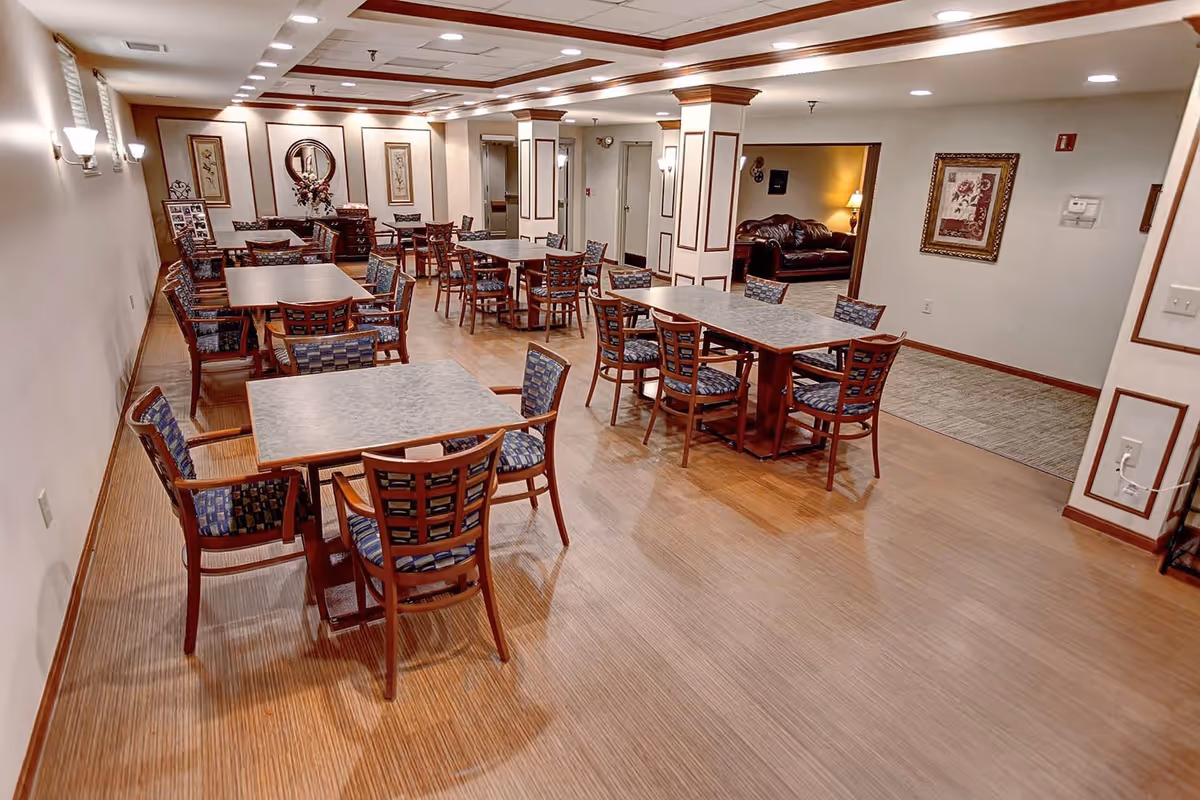 A spacious dining room with multiple tables and chairs arranged neatly on a wooden floor. The room is well-lit with ceiling lights and wall sconces. There are framed pictures and decorative elements on the walls, and an adjacent room with a leather couch is visible in the background.