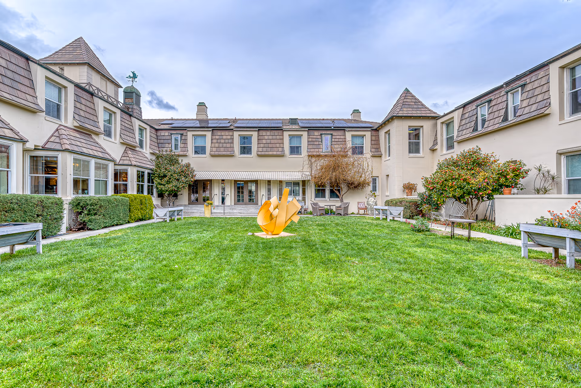 A green courtyard with a central yellow abstract sculpture surrounded by a two-story beige senior living building.