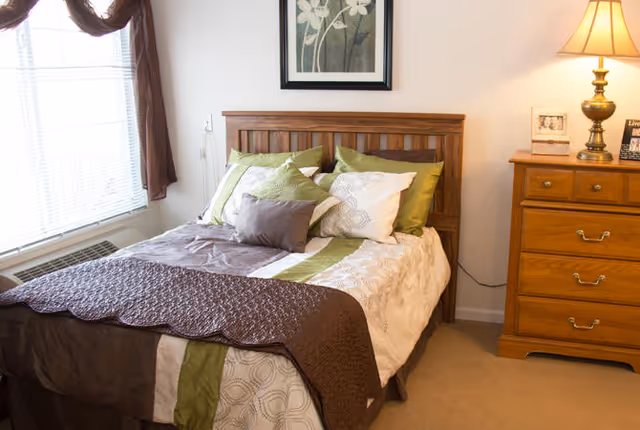 A cozy bedroom with a wooden headboard bed dressed in white, green, and brown bedding with multiple pillows. There is a wooden dresser with a lamp and framed photos on top, and a window with brown curtains letting in natural light.